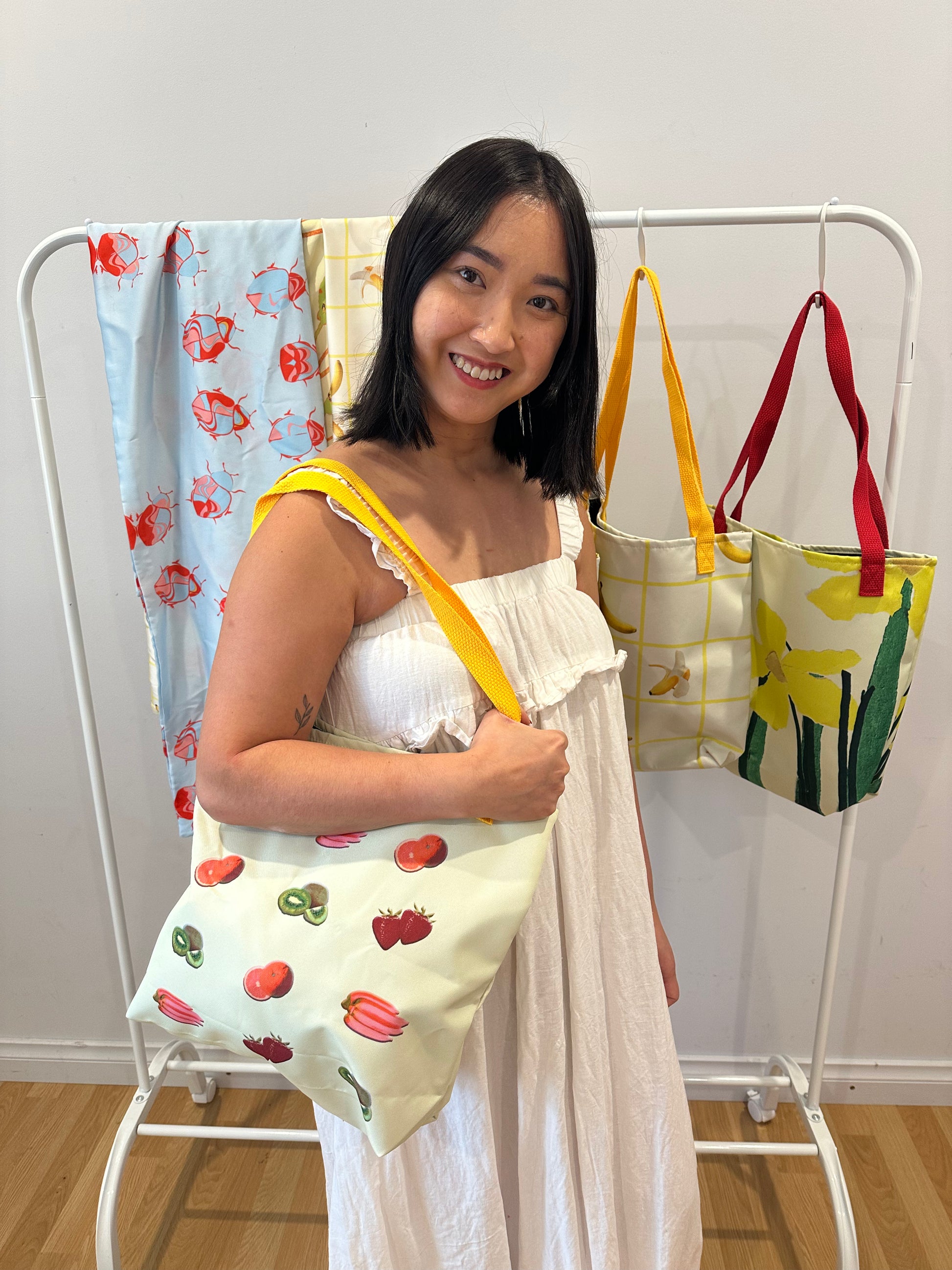 Woman holding Let's Go Shopping tote bag with fruit design in front of a rack with colorful Duyen Del Design products.