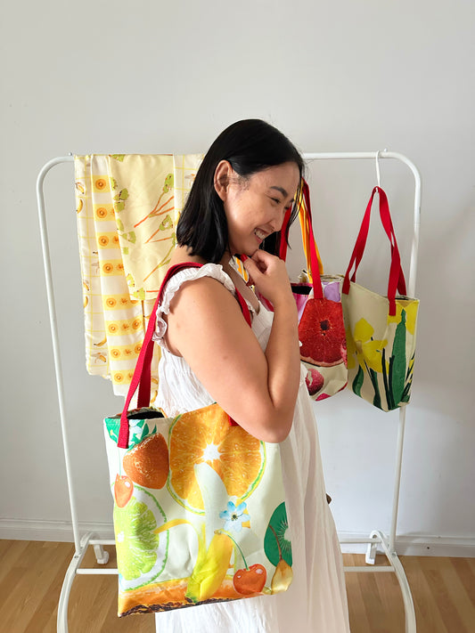 Woman holding the colourful, Juicy Fruits tote bag in front of a rack full of Duyen Del Design products in a room with a white wall and wooden floor.